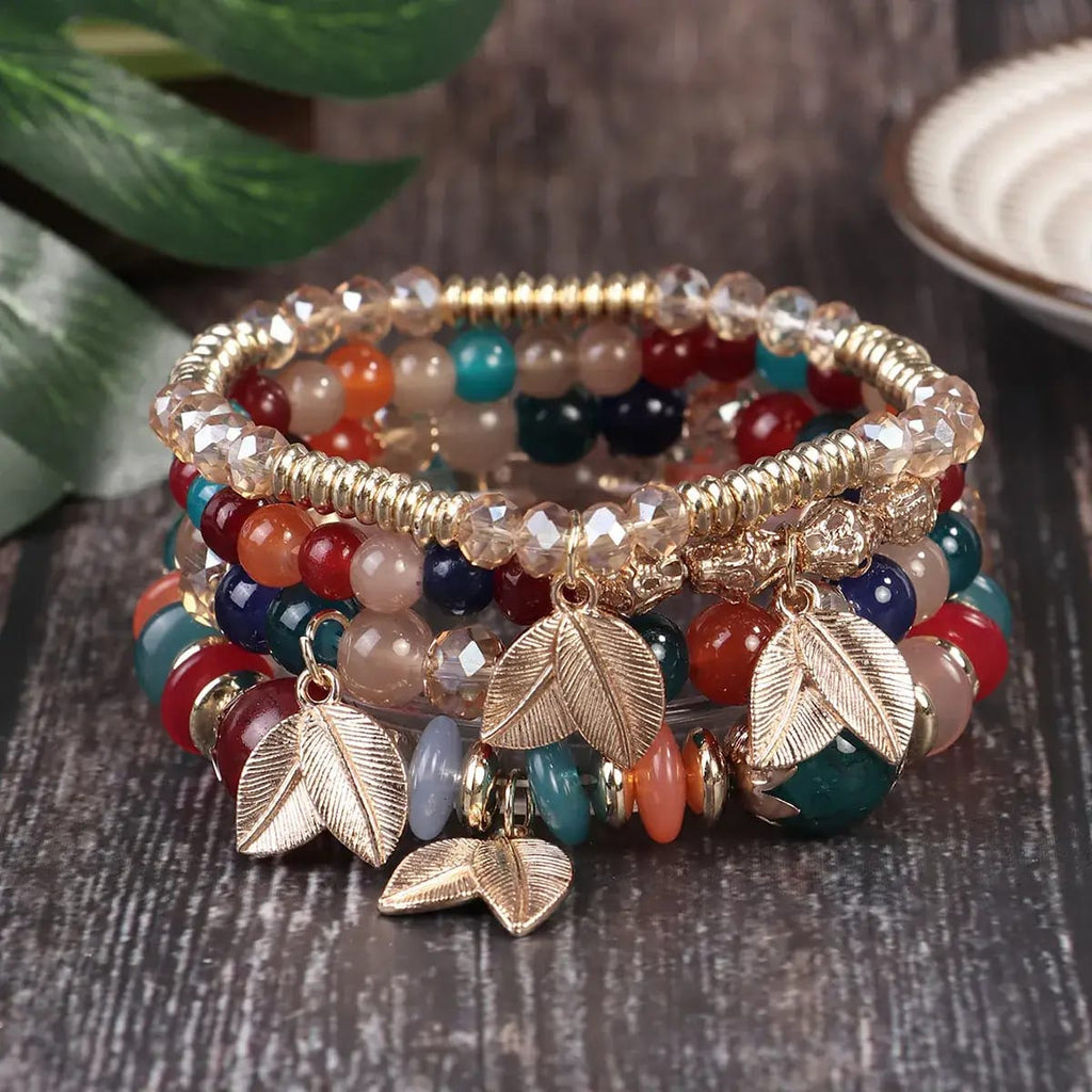 A stack of bohemian beaded bracelets with gold-toned leaf charms, displayed on a dark wooden surface with green leaves and a ceramic plate in the background.