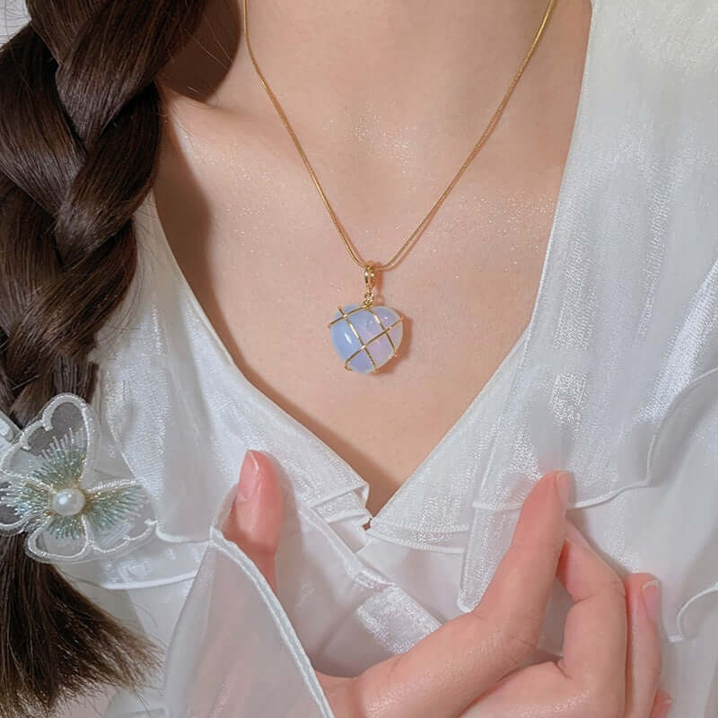 A close-up lifestyle shot of a woman wearing a delicate gold chain necklace with a geometric, faceted pendant made of pale blue and white stones. She is wearing a white lace blouse, and her hair is styled in a braid with a decorative hair clip.