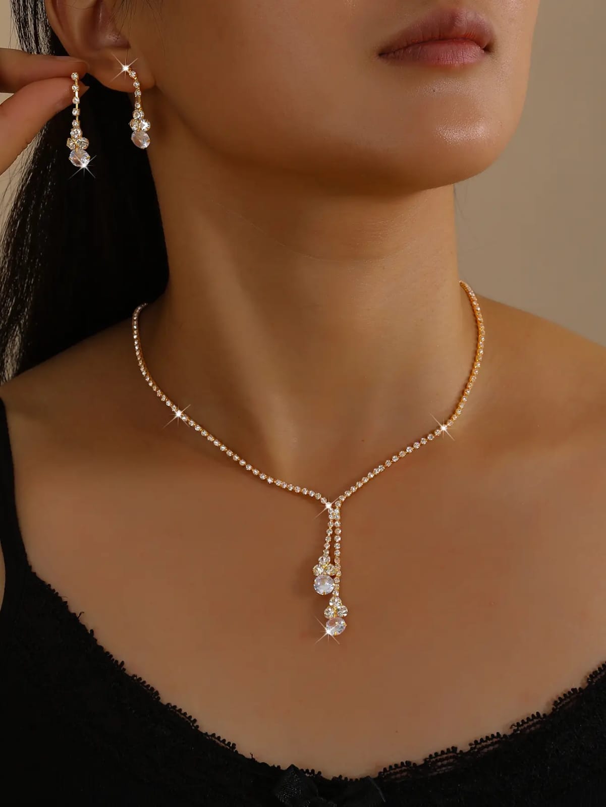 A close-up of a woman wearing a delicate gold necklace with a Y-shaped drop and matching dangling earrings, styled with a black lace top against a warm beige background.