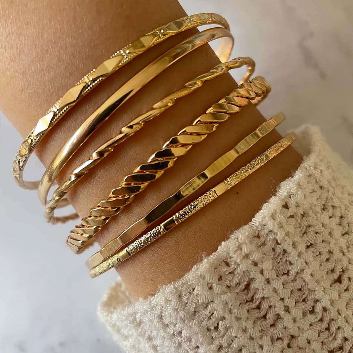 A close-up of a wrist wearing five stacked gold bangle bracelets in different textures, paired with a cream-colored knit sweater cuff.