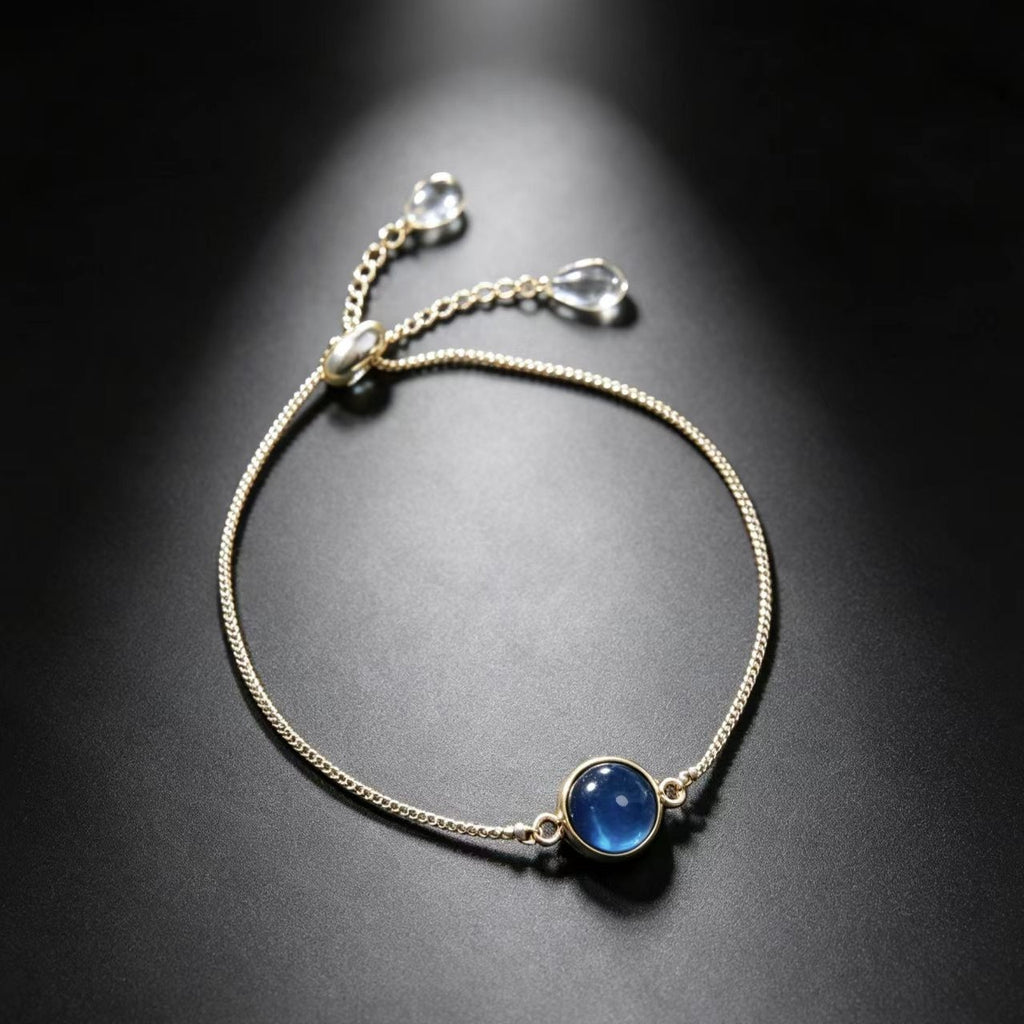 A delicate gold chain bracelet with a round blue cabochon gemstone pendant, displayed on a dark reflective surface with a spotlight effect.