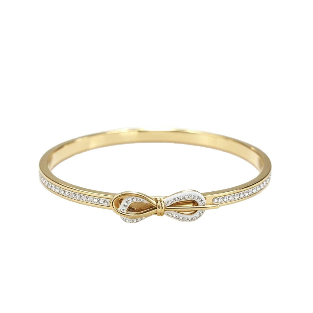 A gold bangle bracelet featuring a central bow motif paved with small clear stones on the front band, shown against a plain white background.