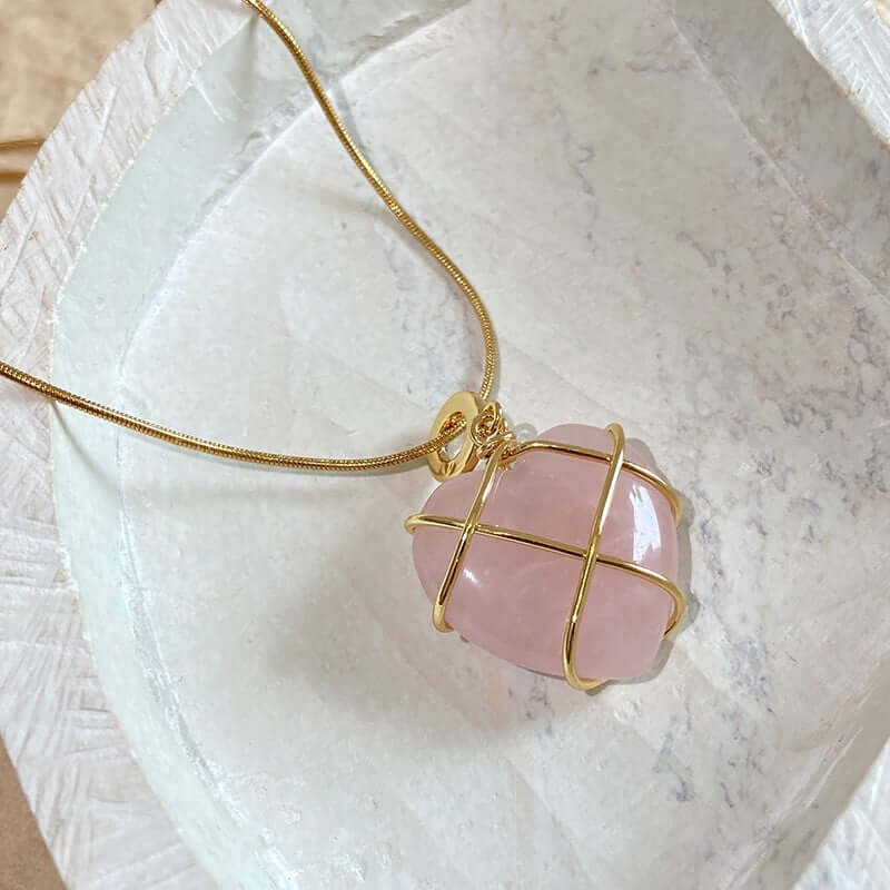 A gold wire cage pendant necklace holding a smooth, polished pink stone, resting on a textured white stone tray.