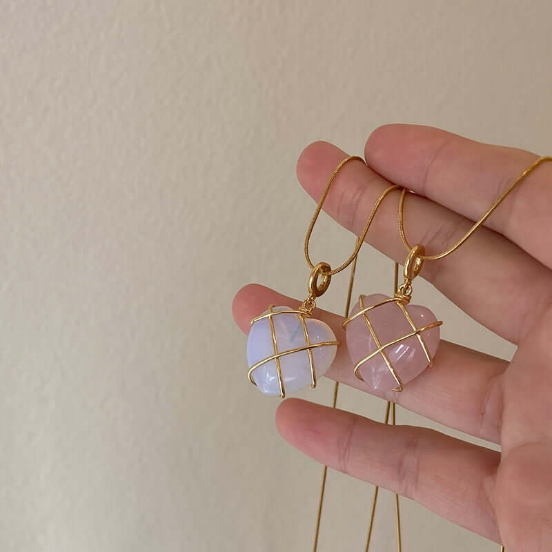 A hand holding two gold wire-wrapped heart-shaped gemstone pendants, one pale white and one soft pink, against a plain beige wall background.