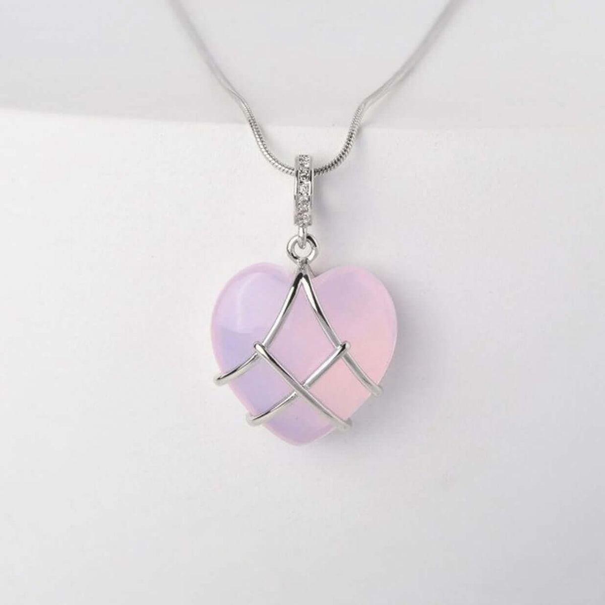A heart-shaped pendant necklace made of iridescent pale pink and lavender stone wrapped in a silver wire pattern, hanging from a silver chain against a white background.