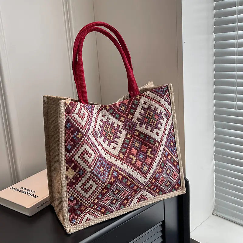 A jute tote bag with a red geometric embroidered pattern and red fabric handles, resting on a dark wooden surface next to a book in a home setting.