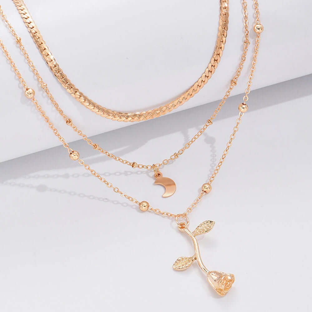 A layered gold necklace set displayed on a white surface, featuring a thick textured chain, a delicate chain with a crescent moon pendant, and a longer chain with a rose-shaped pendant.