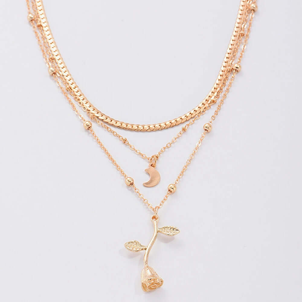 A multi-layered rose gold necklace featuring a textured chain, a beaded chain, and a delicate chain with a crescent moon charm and a rose stem pendant, displayed on a light gray background.