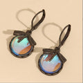 A pair of dangling earrings with dark oxidized metal hooks and oval cabochons showing iridescent blue, purple, and orange reflections, photographed on a plain beige background.