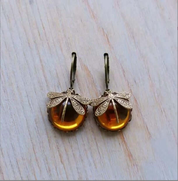A pair of dragonfly-shaped drop earrings with amber-colored oval stones and silver-toned wings, displayed on a light wood surface.