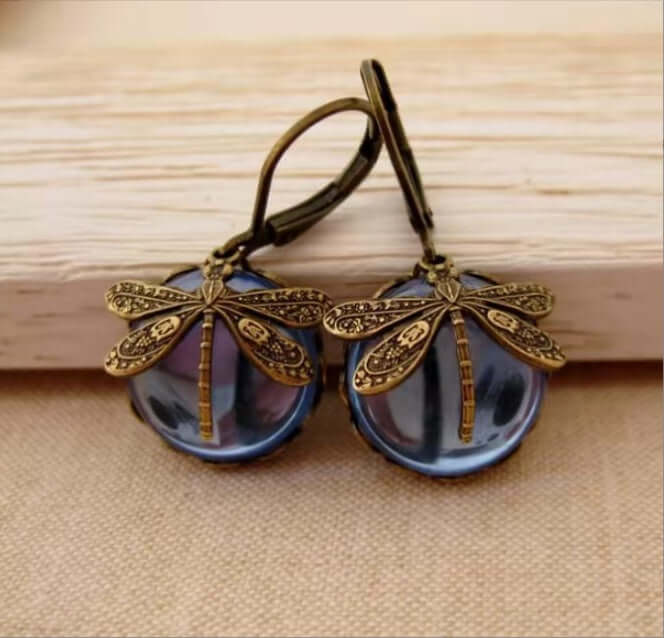 A pair of dragonfly drop earrings with antique bronze metal wings and hooks, hanging in front of oval dark blue iridescent beads on a light wood and fabric surface.