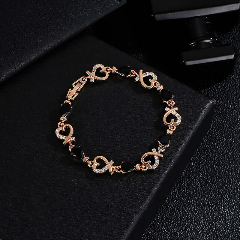 A rose gold bracelet with heart and infinity knot motifs, accented with clear rhinestones and black faceted beads, displayed on a black textured surface.