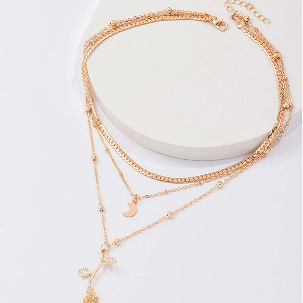 A rose gold multi-layered necklace with a small leaf and flower pendant, displayed on a white circular stand against a light background.