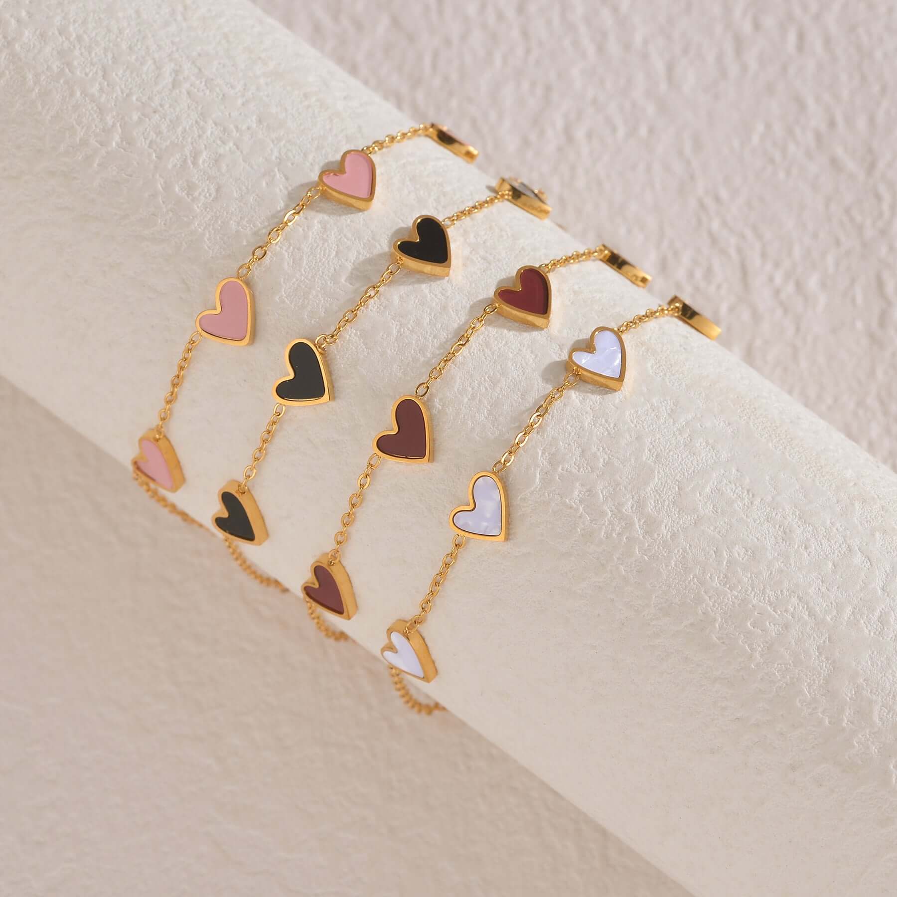 A set of gold chain bracelets with dangling heart charms in pink, black, red, and white, displayed on a textured cream-colored cylindrical stand.