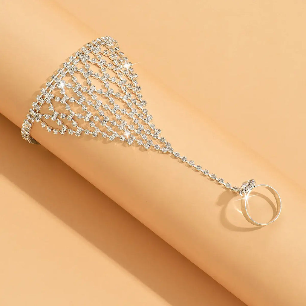 A silver-toned crystal chain bracelet with a dangling ring, displayed on a smooth beige arm against a matching beige background.