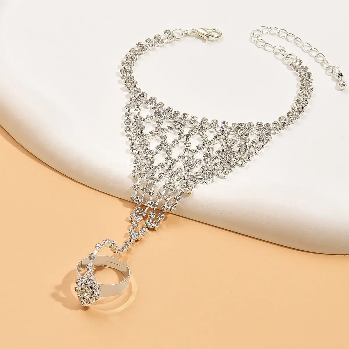 A silver-toned rhinestone choker necklace with a long drop chain ending in a matching rhinestone ring, displayed on a white curved surface over a peach-colored background.