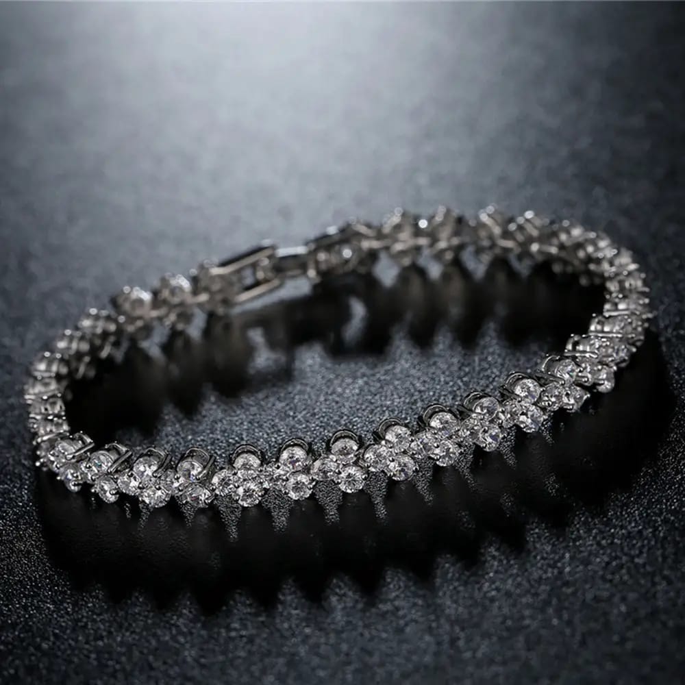 A silver-toned tennis bracelet set with clear round gemstones, displayed on a dark textured surface with a soft reflection.