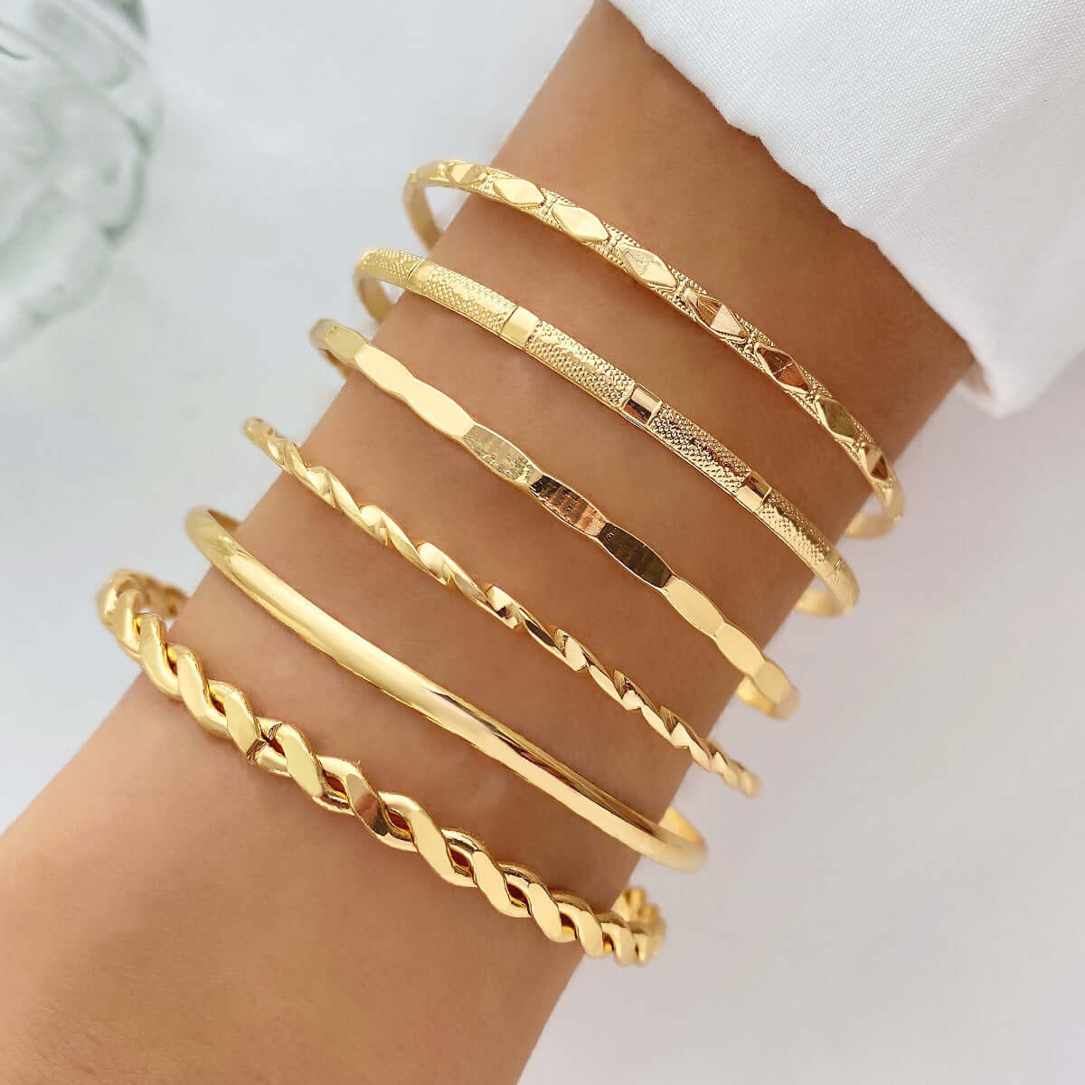A stack of six gold-toned bangle bracelets with different textures and link designs, worn on a person's wrist.