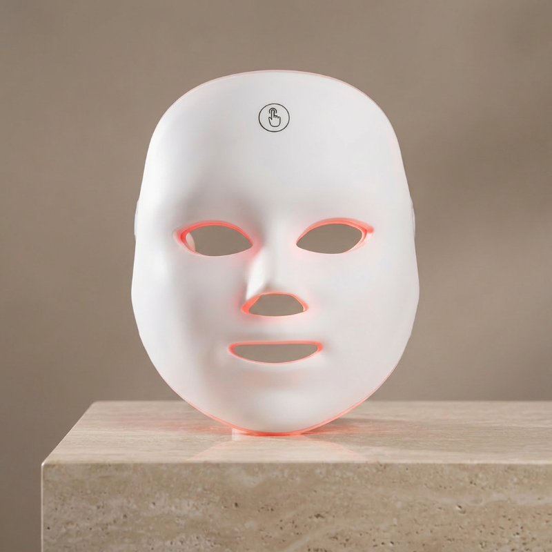 A white LED light therapy face mask resting on a beige stone pedestal against a neutral beige background.