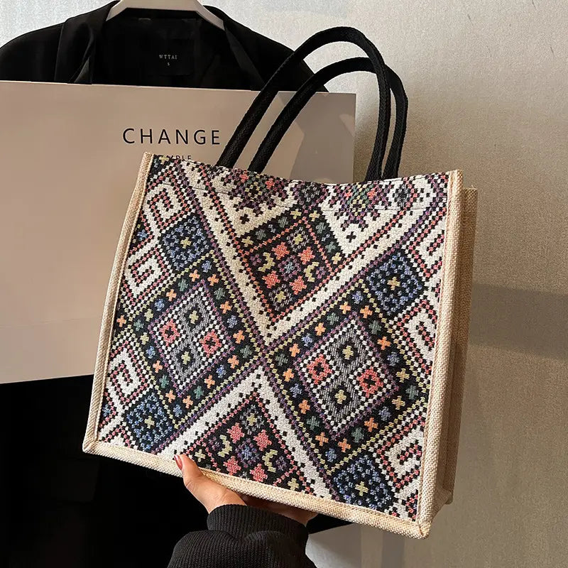 A woven tote bag with a colorful geometric cross-stitch style pattern, held by a hand in front of a black bag and a beige CHANGE shopping bag.