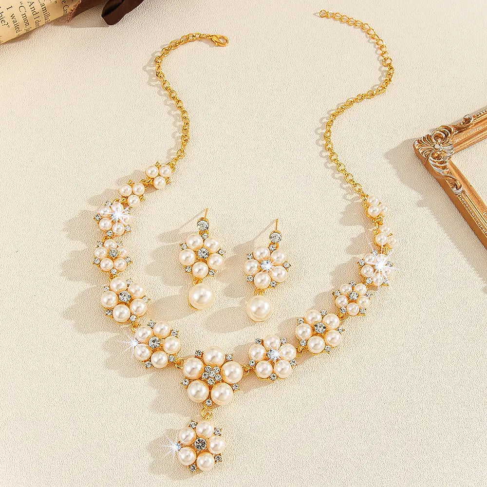 Gold-tone necklace and matching drop earrings set with cream faux pearls and clear rhinestone floral clusters, arranged on a light beige textured surface with decorative props at the corners.