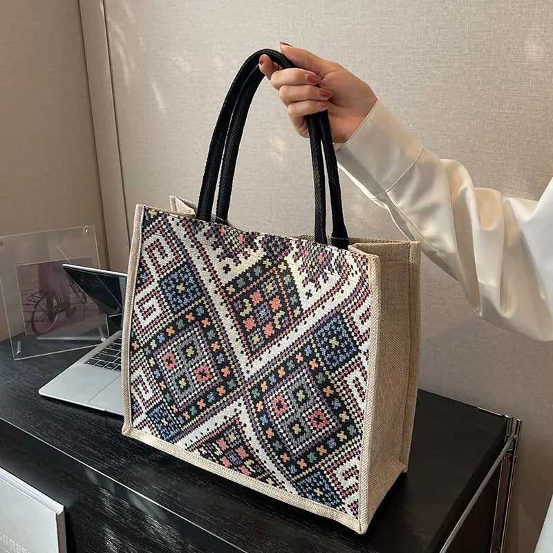 Hand holding a patterned tote bag on a desk with a laptop and photo frame.