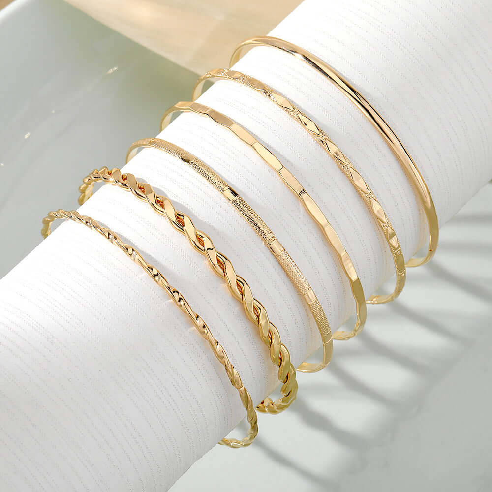 Six gold-toned bangle bracelets in different textures and styles, displayed on a white cylindrical ring stand.