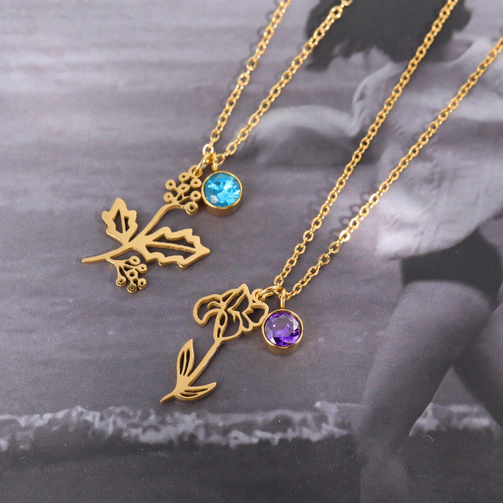 Two gold-tone floral necklaces with round bezel-set gemstones, one light blue and one purple, displayed on a dark, blurred background.
