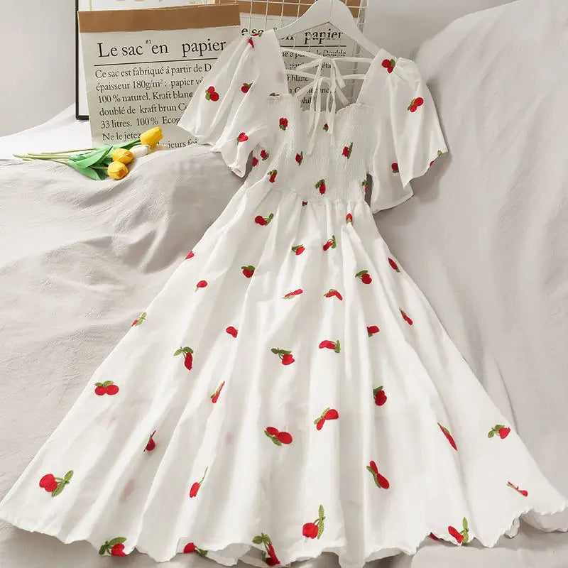 White A-line dress with red strawberry print, featuring short flutter sleeves, a square neckline with tie details, and a gathered waist, displayed on a white fabric surface with decorative props.
