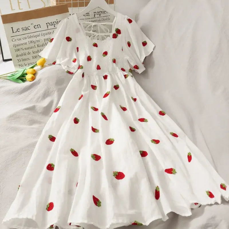 White short-sleeve dress with red strawberry print, featuring a square neckline with lace trim and a gathered waist, displayed on a bed with decorative props.