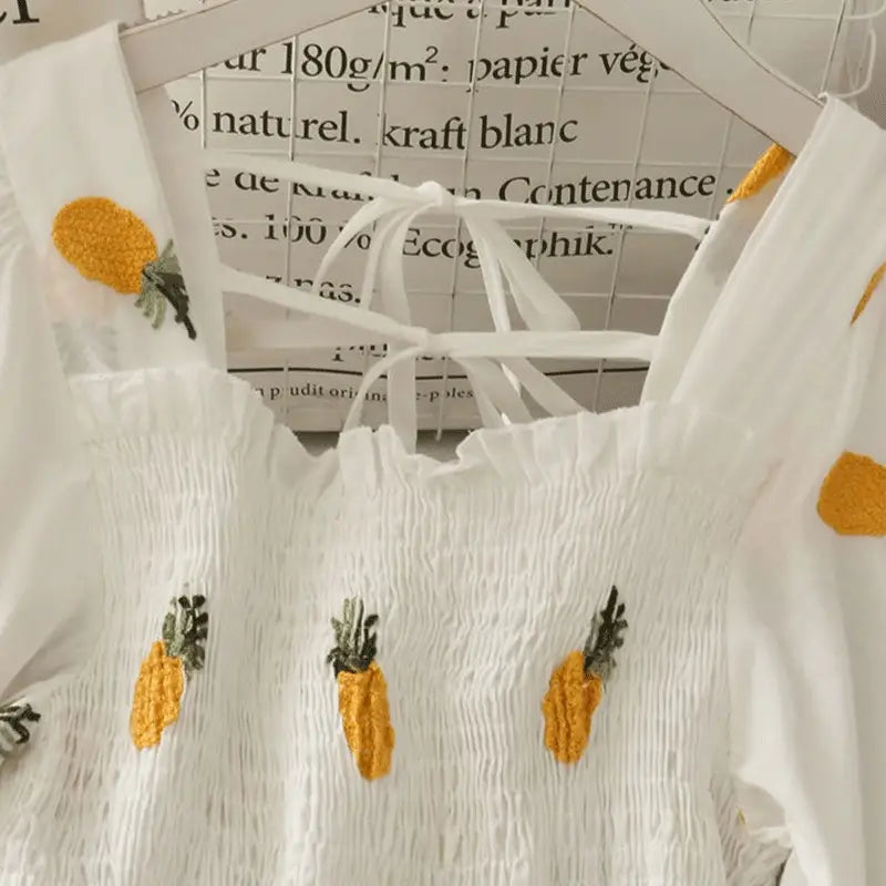 White top with pineapple embroidery on a hanger against a blurred text background