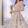 Woman in a floral dress holding flowers by a window with a cityscape view