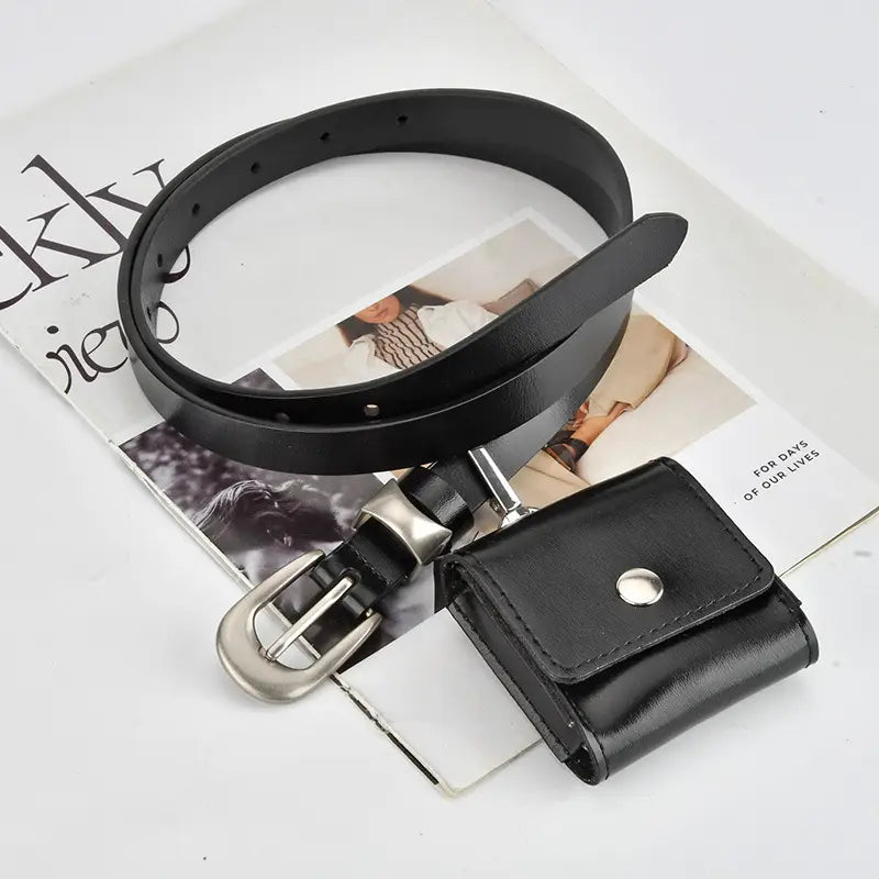 A black leather belt with a silver buckle, shown coiled on top of a magazine page next to a matching black leather card holder pouch.