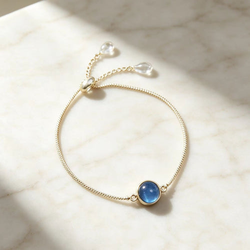 Delicate gold chain adjustable bolo bracelet with round blue crystal charm on light background