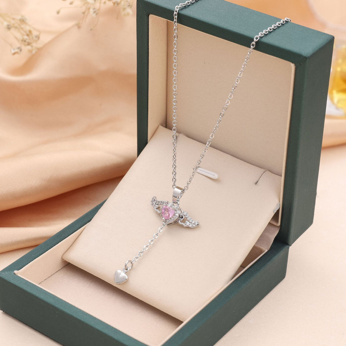 Silver heart wing necklace presented inside a dark green jewelry box on pink satin.