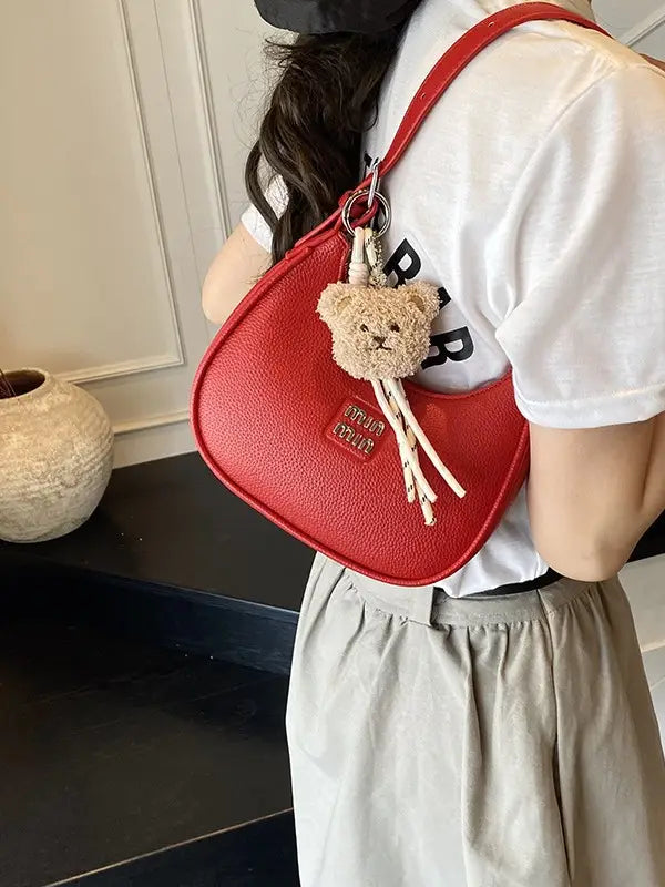​ Woman wearing a vibrant red sparkly crescent hobo shoulder bag with a plush brown teddy bear charm hanging from the strap
