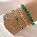 Four layered gold bracelets worn on a wrist, featuring a snake wavy chain, an emerald green gem chain, a thin cuff, and a green beaded strand.