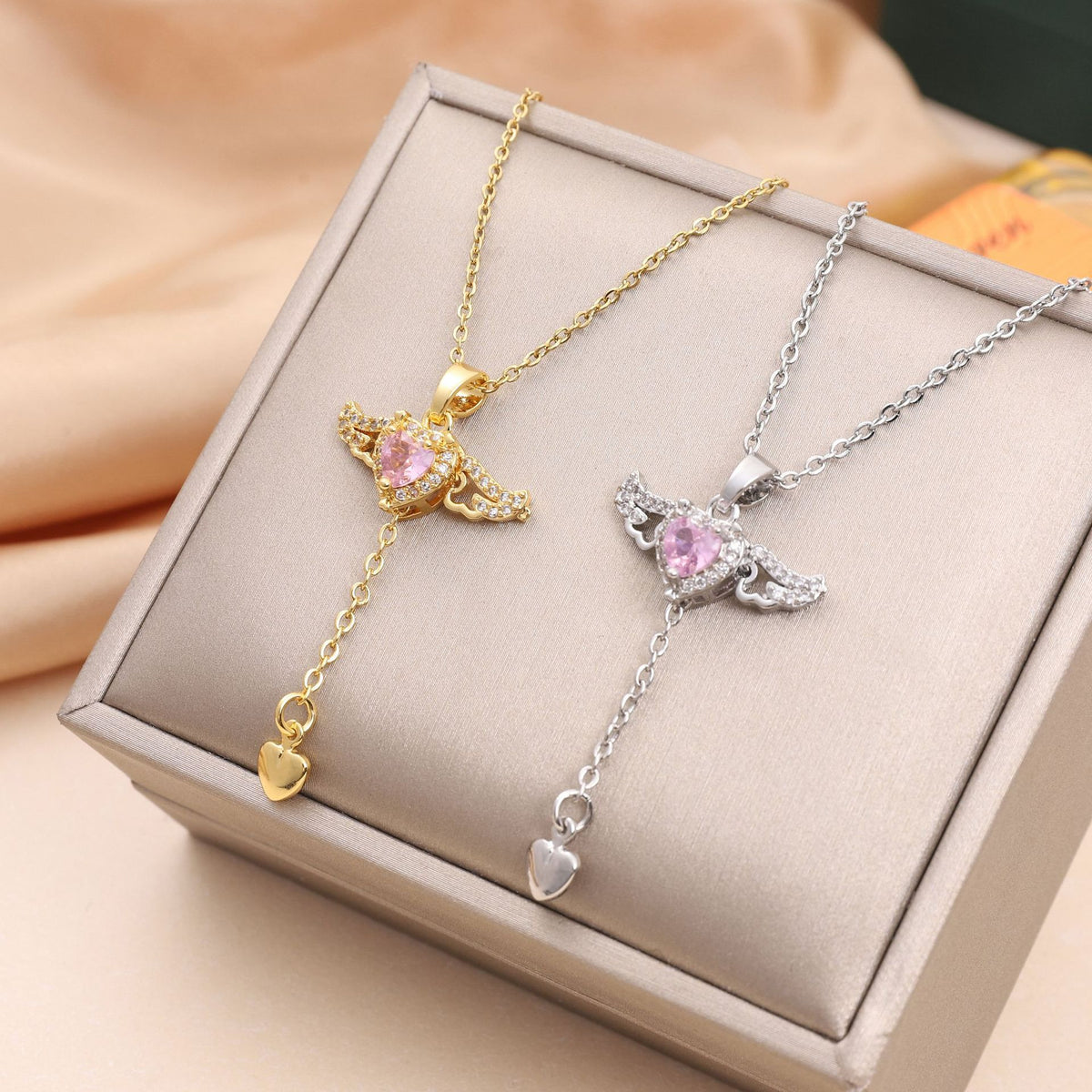Gold and silver angel wing pendant necklaces with pink heart crystals draped over a luxury gray jewelry box.