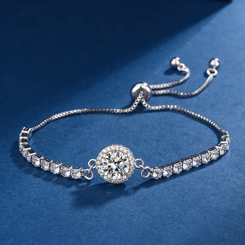 A silver-tone adjustable bracelet featuring a central round halo of clear cubic zirconia stones, flanked by smaller matching stones on each side, displayed on a textured dark blue background.
