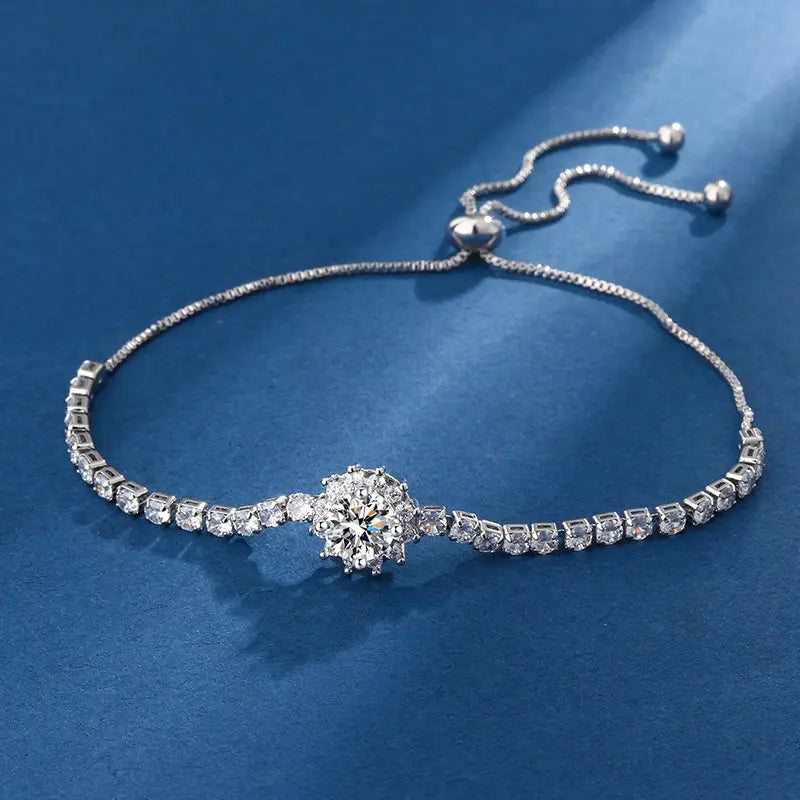 A silver-toned adjustable bracelet featuring a central round-cut clear gemstone surrounded by smaller stones, with a line of small square-cut stones along the band, displayed on a textured blue background.