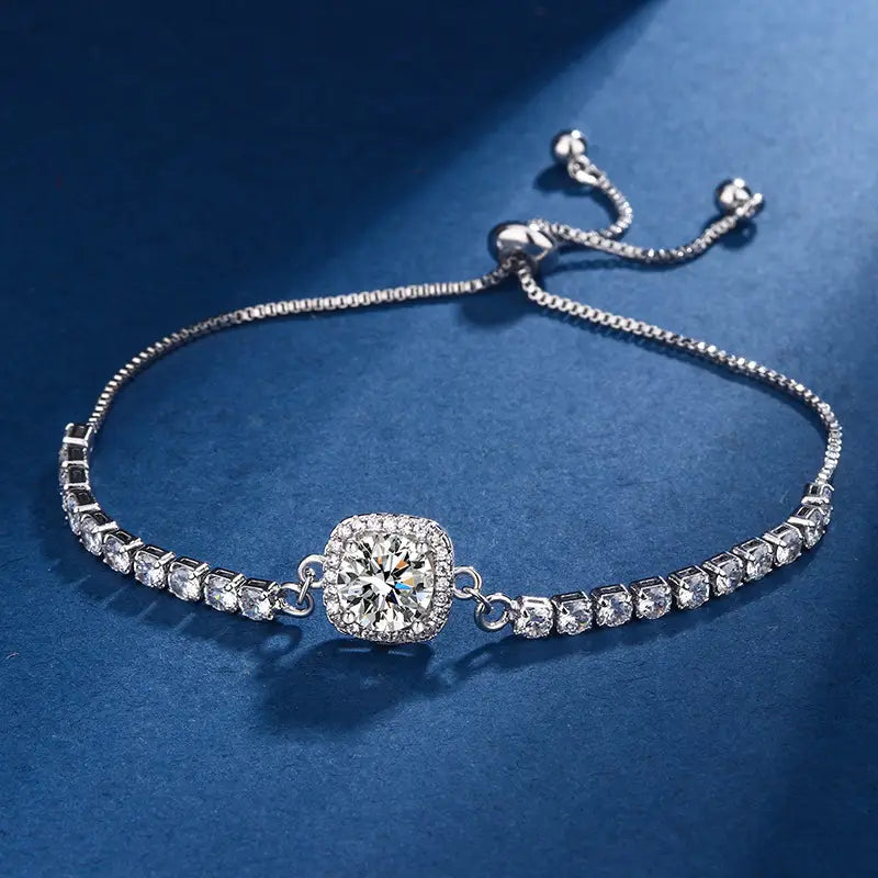 A silver-toned adjustable bracelet featuring a central square halo of clear stones and a row of smaller stones along the band, displayed on a dark blue textured background.