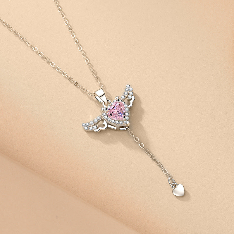 Silver y-lariat necklace with pink heart crystal and spread angel wings on a beige silk surface.